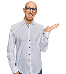 Bald man with beard wearing business shirt and glasses smiling cheerful presenting and pointing with palm of hand looking at the camera.