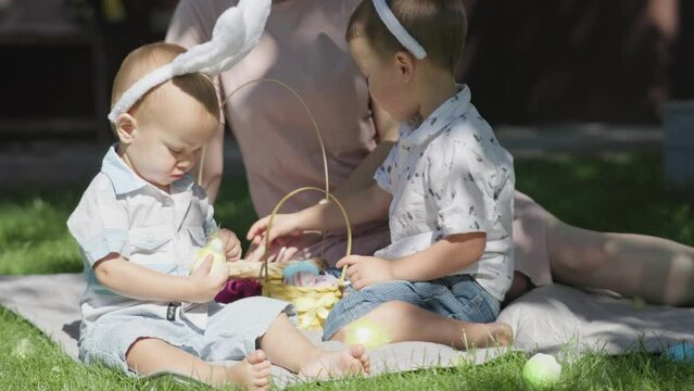 Easter holiday outdoor family mother children sitting spring grass having Easter picnic in park backyard sunny day. Children playing colorful Easter eggs, kids wearing bunny ears having fun together