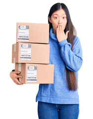 Young beautiful chinese woman holding delivery cardboard boxes covering mouth with hand, shocked and afraid for mistake. surprised expression