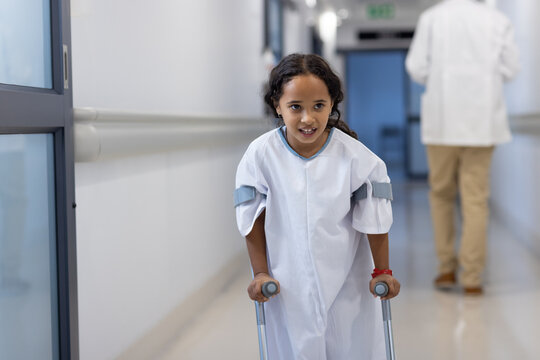 Happy biracial girl walking on crutsches in hospital corridor