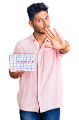 Handsome latin american young man holding heart calendar with open hand doing stop sign with serious and confident expression, defense gesture