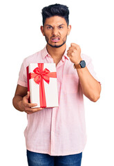 Handsome latin american young man holding gift annoyed and frustrated shouting with anger, yelling crazy with anger and hand raised