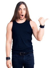 Young adult man with long hair wearing goth style with black clothes surprised pointing with hand...