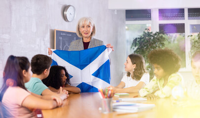 Fototapeta premium Friendly high school woman teacher tells pupils the history of Scotland in class and holds the national flag of the country .in her hands