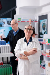 Elderly drugstore worker in white coat holding cup of coffee looking at camera while working in pharmacy, helping clients with health care information. Medicine service and support