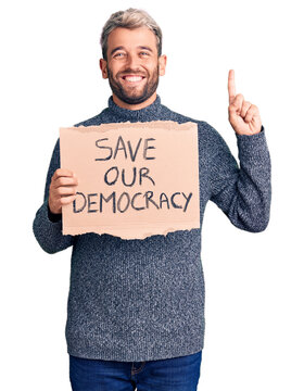Young blond man holding save our democracy cardboard banner surprised with an idea or question pointing finger with happy face, number one