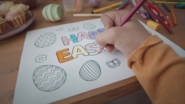 POV of little girl using color pencils while drawing beautiful Easter postcard sitting at kitchen table together with family having preparations