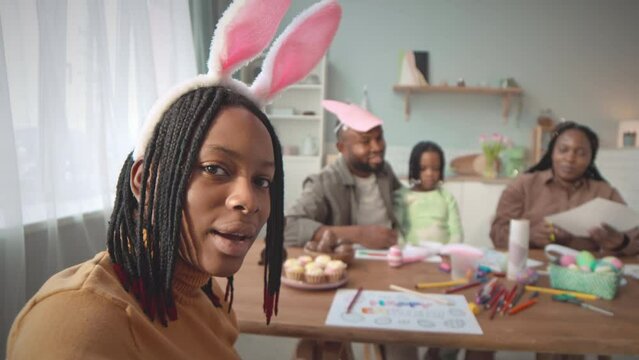 Handheld POV shot of African American teenage girl wearing funny bunny ears on her head recording video of herself making diy Easter decorations together with her parents and little sister