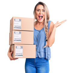 Young beautiful blonde woman holding delivery boxes celebrating victory with happy smile and winner expression with raised hands