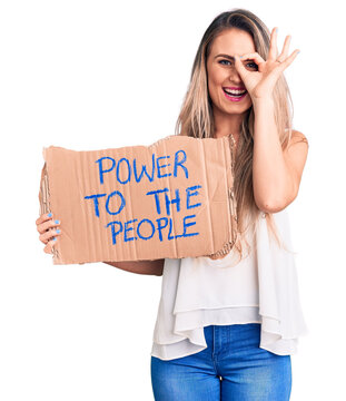 Young beautiful blonde woman holding power to the people cardboard banner smiling happy doing ok sign with hand on eye looking through fingers
