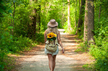 A blonde caucasian woman walks down a trail in the forest with sunflowers sticking out of her backpack on a sunny Summer's Day  - Powered by Adobe