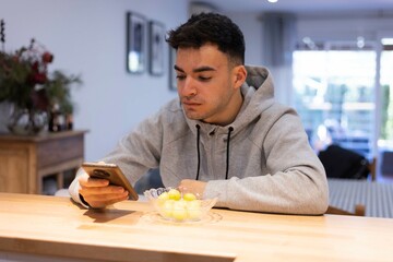 Hombre joven esta mirando el móvil mientras desayuna unas uvas.