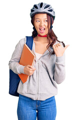 Young beautiful chinese girl wearing student backpack and bike helmet holding book pointing thumb up to the side smiling happy with open mouth