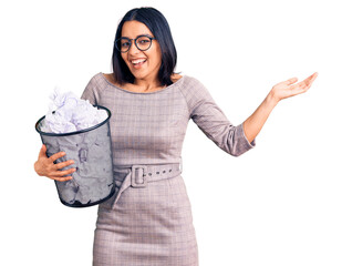 Young beautiful latin woman holding paper bin full of crumpled papers celebrating victory with happy smile and winner expression with raised hands