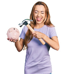 Young caucasian woman holding piggy bank and hammer sticking tongue out happy with funny expression.