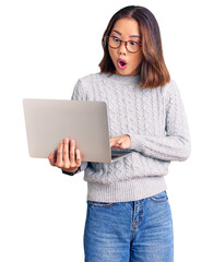 Young beautiful chinese girl wearing glasses holding laptop scared and amazed with open mouth for...