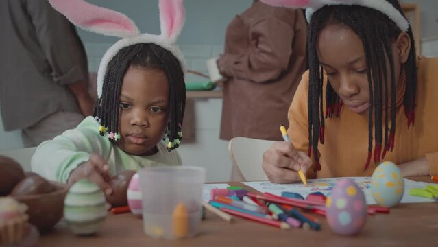 Waist up of two African American girls in cute bunny ears headbands sitting at kitchen table together making DIY Easter decorations