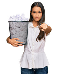 Beautiful hispanic woman holding paper bin full of crumpled papers pointing with finger to the camera and to you, confident gesture looking serious