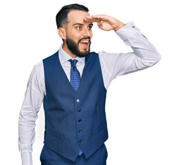 Young man with beard wearing business vest very happy and smiling looking far away with hand over head. searching concept.