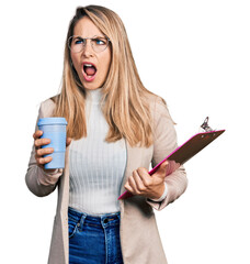 Young blonde woman wearing business style, drinking coffee and holding clipboard angry and mad...