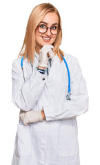 Beautiful caucasian woman wearing doctor uniform and stethoscope looking confident at the camera with smile with crossed arms and hand raised on chin. thinking positive.