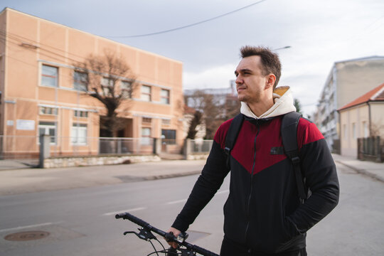 A young guy is going through the city while pushing his bicycle during the day