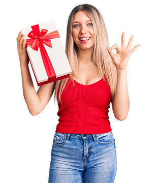 Young beautiful blonde woman holding gift doing ok sign with fingers, smiling friendly gesturing excellent symbol