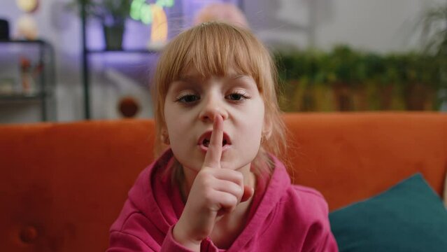 Shh be quiet please. Young blonde child kid girl presses index finger to lips makes silence hush gesture sign do not tell gossip privacy secret. Female teen toddler at home in living room sits on sofa