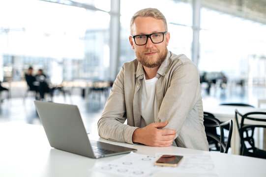 Clever successful caucasian man, with glasses, in stylish casual wear, IT specialist, company seo, programmer, digital developer, sits at a desk with a laptop in coworking, looks at camera, smiles