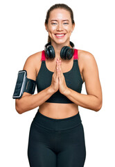 Beautiful blonde woman wearing gym clothes and using headphones praying with hands together asking for forgiveness smiling confident.