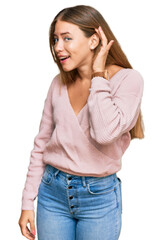 Beautiful blonde woman wearing casual winter pink sweater smiling with hand over ear listening an hearing to rumor or gossip. deafness concept.