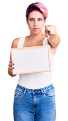 Young beautiful woman with pink hair holding empty white chalkboard pointing with finger to the camera and to you, confident gesture looking serious