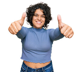 Young hispanic woman with curly hair wearing casual clothes approving doing positive gesture with hand, thumbs up smiling and happy for success. winner gesture.