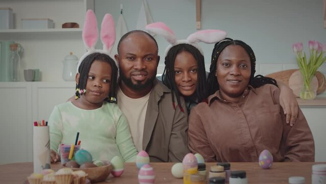 Zoom portrait of cheerful African American family with two kids posing for camera in bright cozy kitchen with Easter eggs, crafts and decoration on wooden table