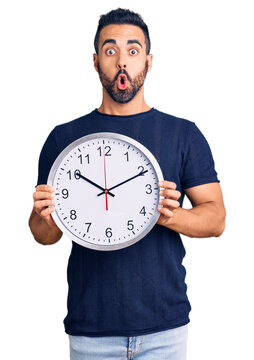 Young hispanic man holding big clock scared and amazed with open mouth for surprise, disbelief face
