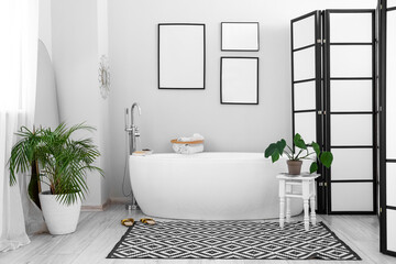 Fototapeta premium Interior of light bathroom with bathtub, blank frames and houseplants
