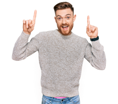 Young redhead man wearing casual winter sweater smiling amazed and surprised and pointing up with fingers and raised arms.