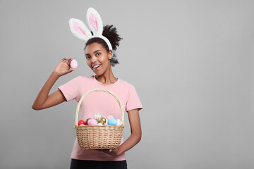 Happy African American woman in bunny ears headband holding wicker basket with Easter eggs on gray...