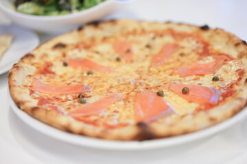 Delicious hot pizza with smoked salmon, on wooden table ready to eat. Smoked salmon pizza with salmon and mozzarella. Smoked salmon pizza front of blurry green salad background.