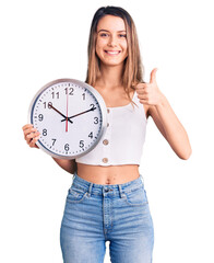 Young beautiful girl holding big clock smiling happy and positive, thumb up doing excellent and approval sign