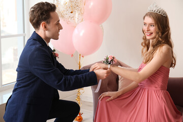 Teenage boy tying corsage around his girlfriend's wrist for prom in room
