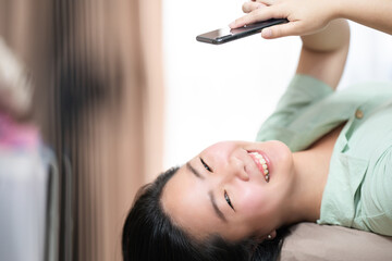 Fototapeta premium Beautiful Asian woman is lying down and playing mobile on the bed in her bedroom with a pastel green - brown color theme.