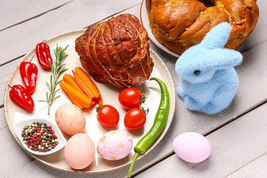 Plate with tasty ham, painted Easter eggs, vegetables and bunny on light wooden background, closeup