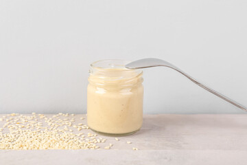 Glass jar of tasty tahini, sesame seeds and spoon on light table