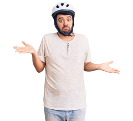 Young hispanic man wearing bike helmet clueless and confused expression with arms and hands raised. doubt concept.