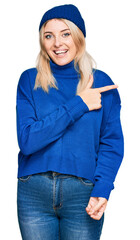 Young caucasian woman wearing wool winter sweater and cap cheerful with a smile of face pointing with hand and finger up to the side with happy and natural expression on face