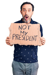 Middle age handsome man holding not my president carboard banner scared and amazed with open mouth for surprise, disbelief face