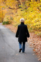  Mature blond woman walking down forest path admiring the colorful Autumn leaves.