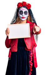 Woman wearing day of the dead costume holding empty white chalkboard pointing with finger to the camera and to you, confident gesture looking serious