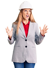 Beautiful young woman wearing architect hardhat crazy and mad shouting and yelling with aggressive expression and arms raised. frustration concept.